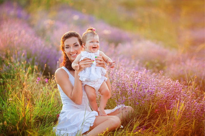 Woman wearing white dress holding up her infant daughter in a field of lavender.