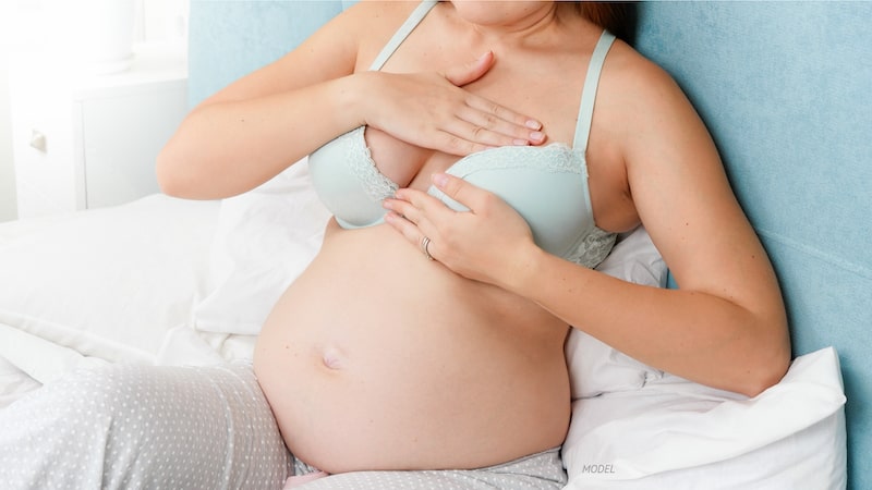 Pregnant woman sitting on bed, feeling top of her breast.
